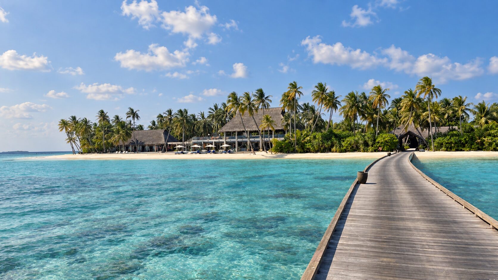 A wooden jetty leading across a turquoise lagoon to a small palm-covered island in the Maldives.